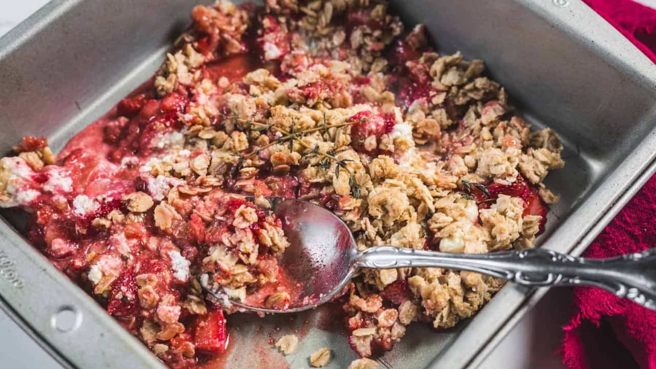 A serving of fruit crumble with a crumbly oat topping and a layer of red fruit filling, garnished with a sprig of thyme on a white plate with a brown rim, next to a silver spoon.