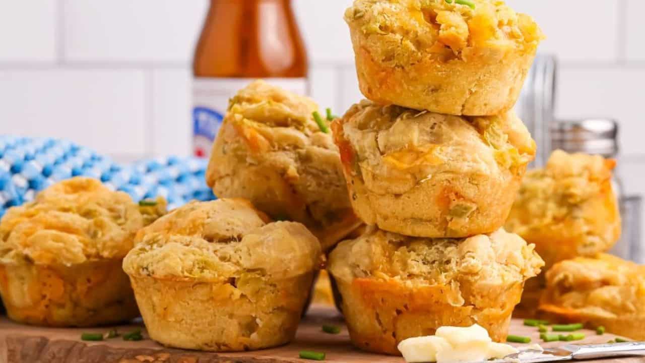 A stack of savory muffins with herbs and cheese sits on a wooden board, with butter, chives, a bottle, and kitchen items in the background.