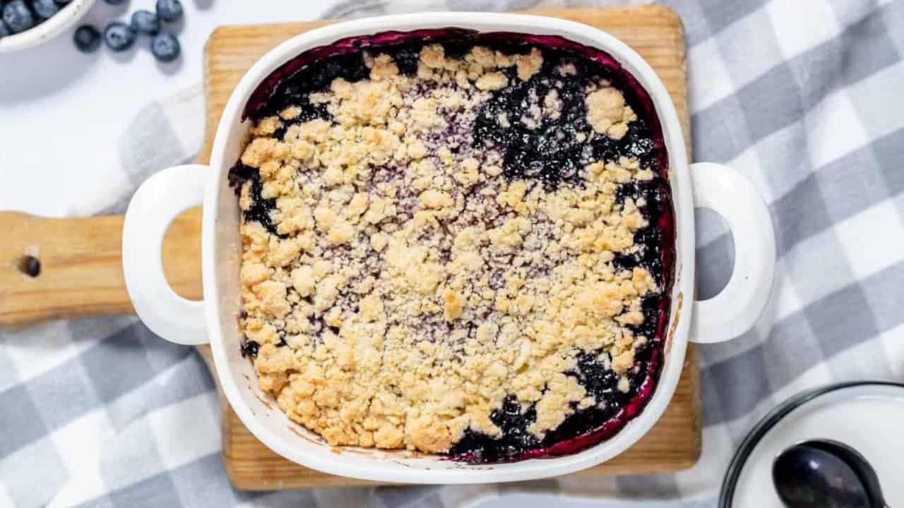 A square baking dish filled with blueberry crisp topped with a crumbly golden brown streusel, placed on a wooden board with a blue cloth and scattered blueberries beside it.
