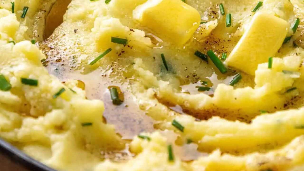 The image shows a bowl of mashed potatoes topped with pepper and chopped chives, resting on a plaid cloth.