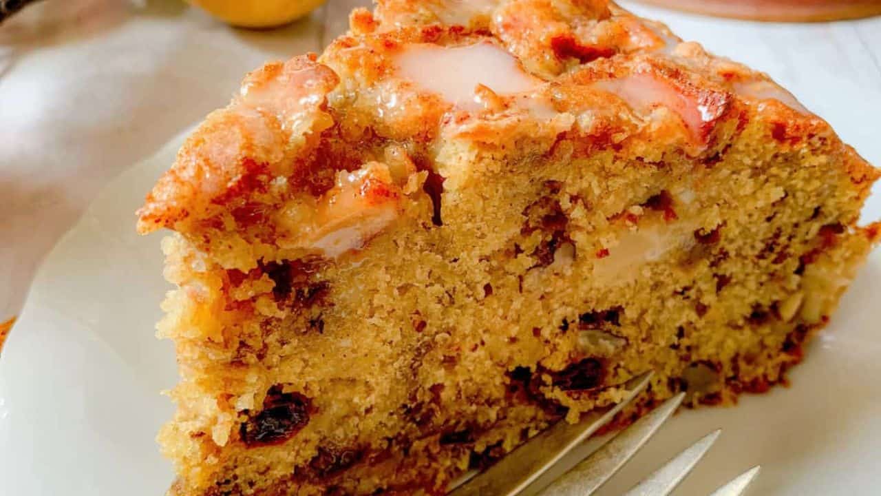 A slice of fruit cake with visible nuts and dried fruits on a white plate, accompanied by a fork.
