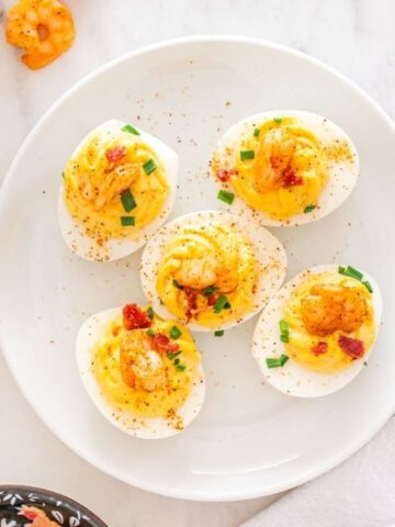 A plate of deviled eggs topped with shrimp and garnished with chopped herbs, next to a bowl of salt and a white napkin.