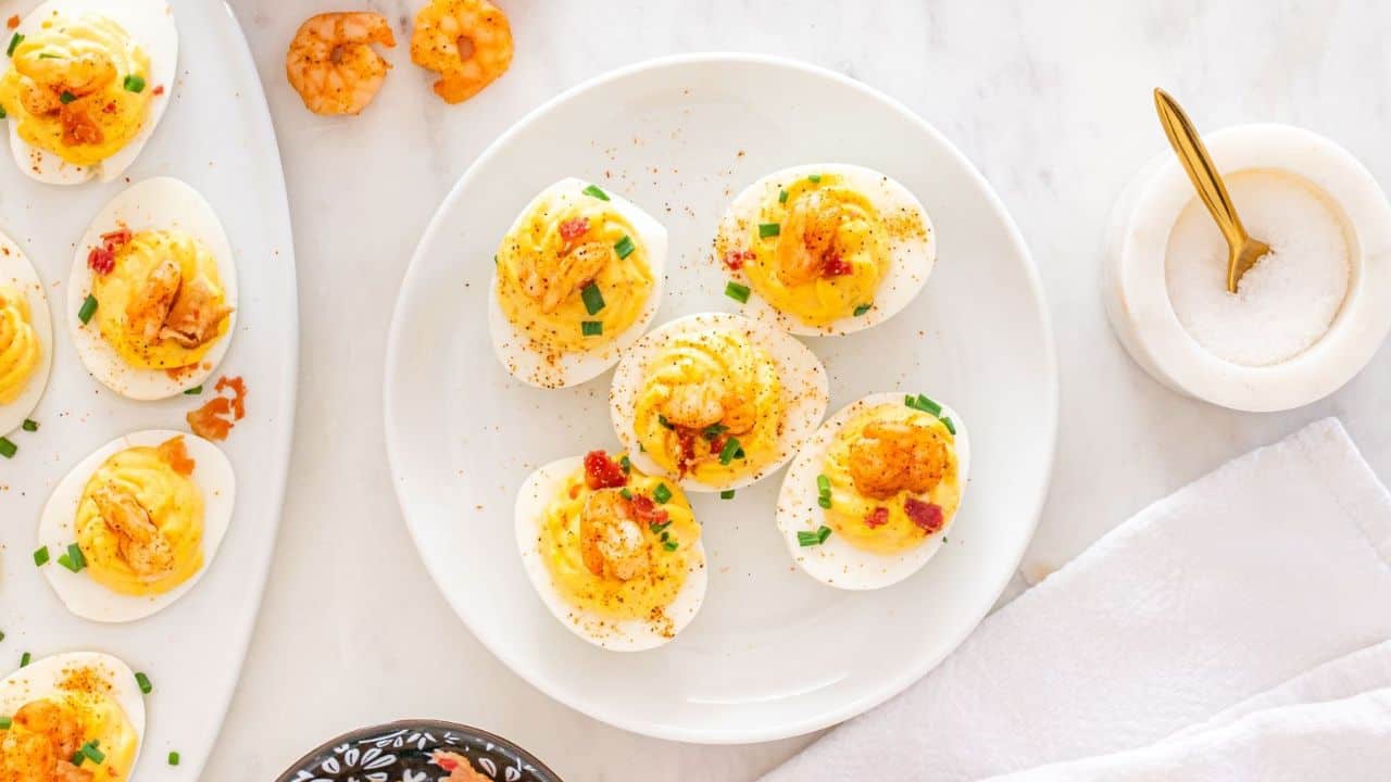 A plate of deviled eggs topped with shrimp and garnished with chopped herbs, next to a bowl of salt and a white napkin.
