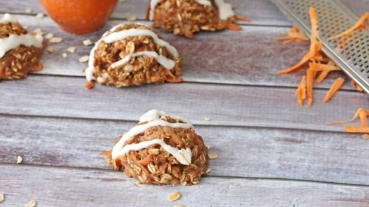 A no-bake oatmeal and carrot cookie topped with white icing sits on a wooden surface, with grated carrot and a metal grater nearby.