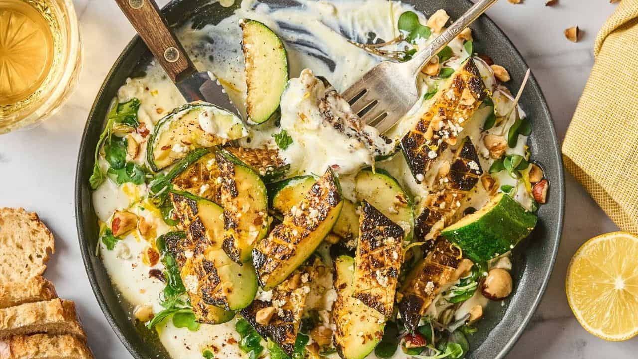 Grilled zucchini slices on a plate with creamy sauce, greens, chopped nuts, and a fork; bread, lemon, and a glass of drink are nearby.