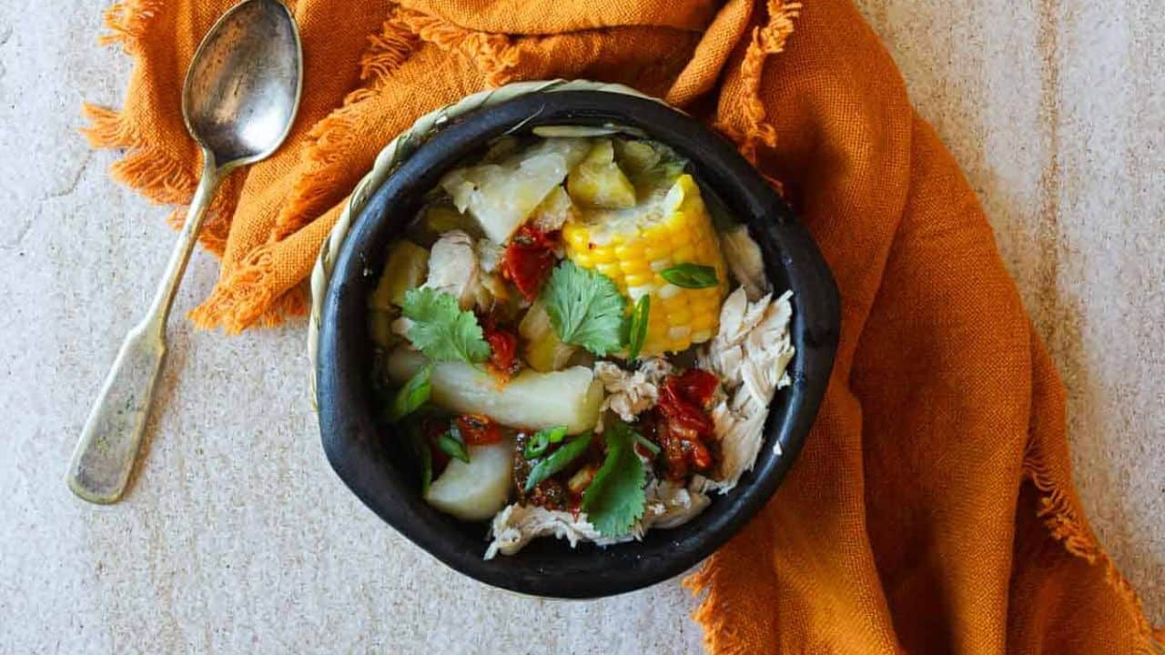 Bowl of chicken sancocho with corn, plantains, potatoes, and carrots in broth.