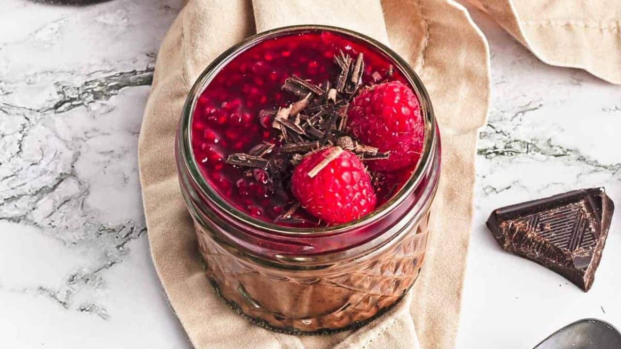 Two small jars filled with chocolate mousse, topped with raspberry sauce, fresh raspberries, and chocolate shavings. A beige cloth, chocolate pieces, loose raspberries, and a spoon are nearby on a marble surface.
