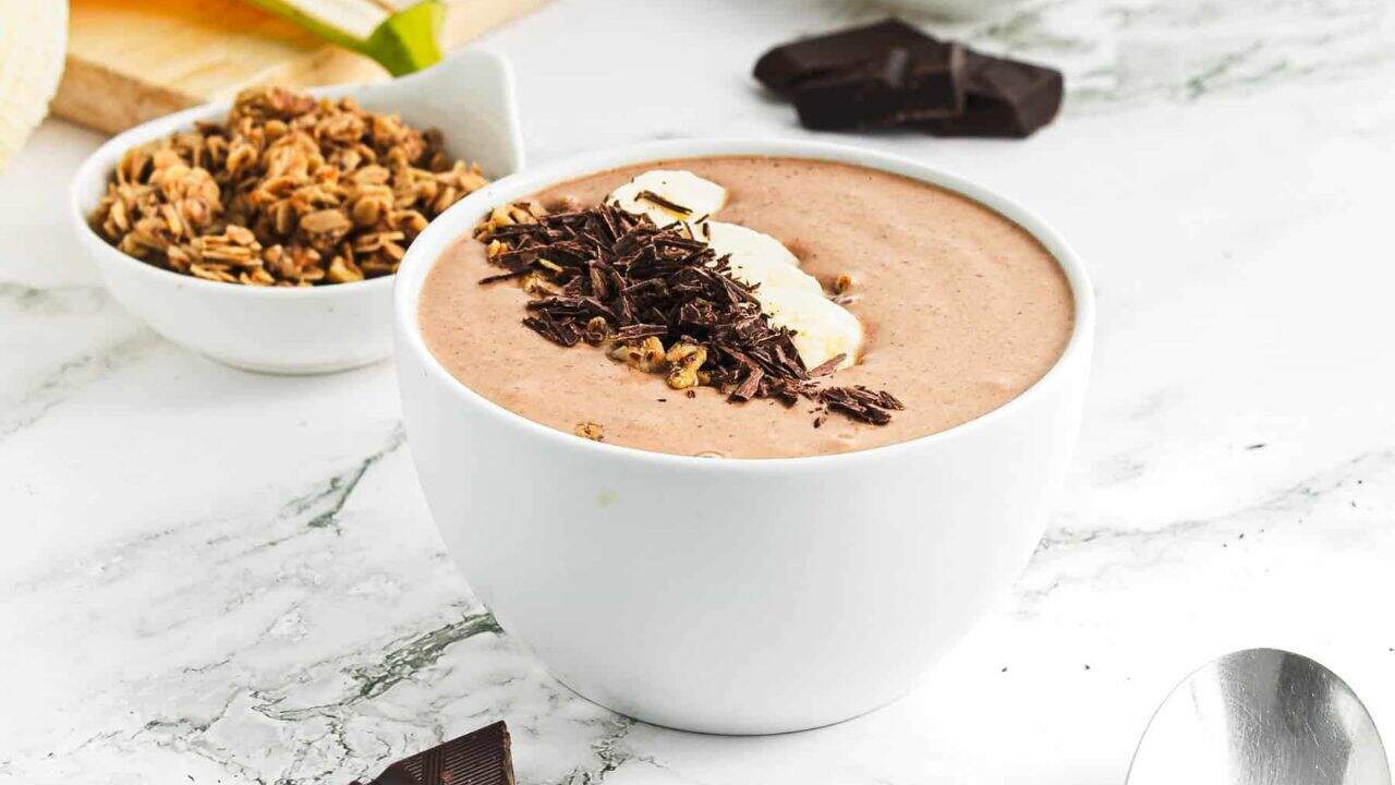 A bowl filled with a chocolate smoothie, topped with banana slices, granola, and chocolate shavings, sits on a white marble surface next to a spoon and a piece of chocolate.