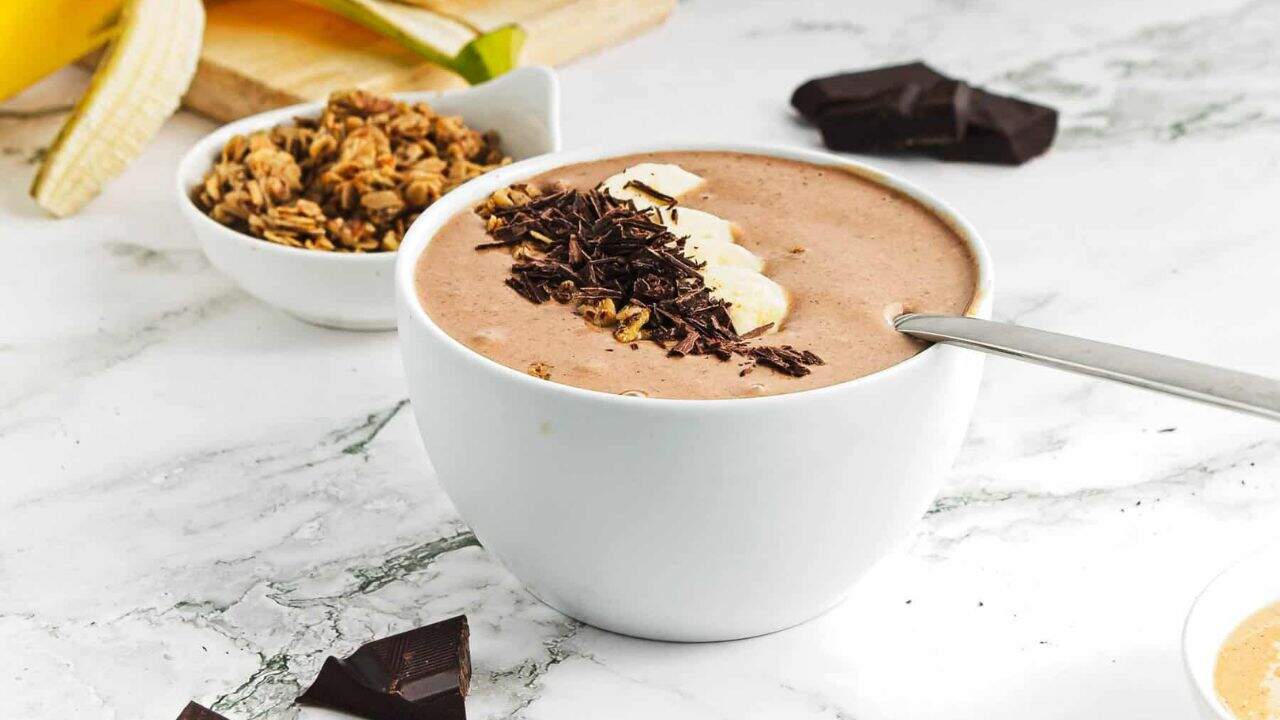 A bowl filled with a chocolate smoothie, topped with banana slices, granola, and chocolate shavings, sits on a white marble surface next to a spoon and a piece of chocolate.