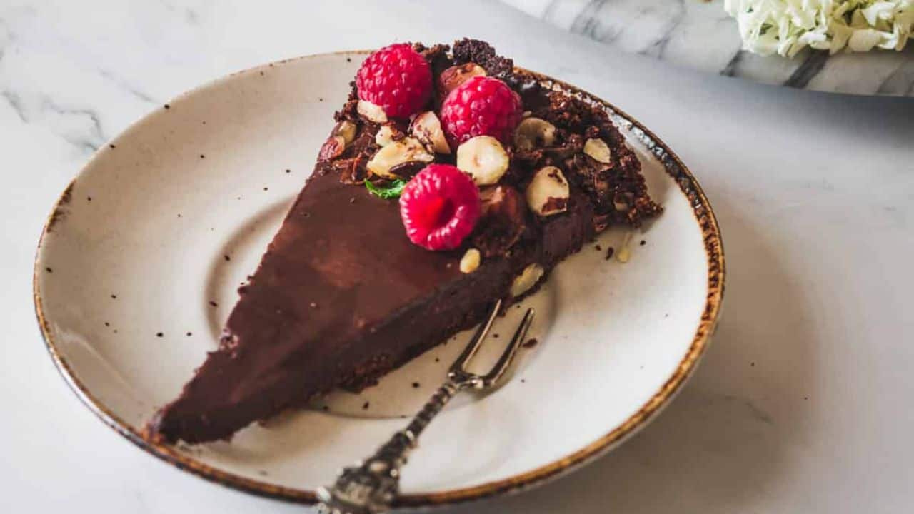 A slice of chocolate tart topped with raspberries and chopped nuts is served on a plate with a small dessert fork.