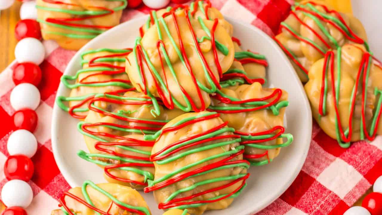 A plate of holiday-themed cookies is placed on a red and white checkered cloth. The cookies are drizzled with red and green icing over a caramel-colored base, surrounded by red and white candy beads.