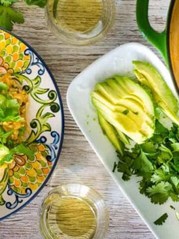 A plate of rice and chicken topped with avocado and cilantro, next to a dish of sliced avocado, lime wedges, cilantro, and a pot of rice and chicken. Glasses of water are also visible.