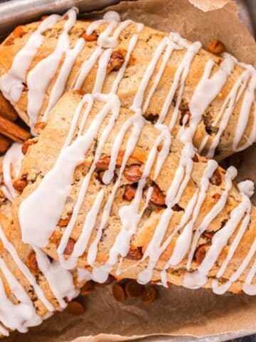 Three glazed scones with cinnamon chips on parchment paper, next to a bundle of cinnamon sticks and a small white pitcher.