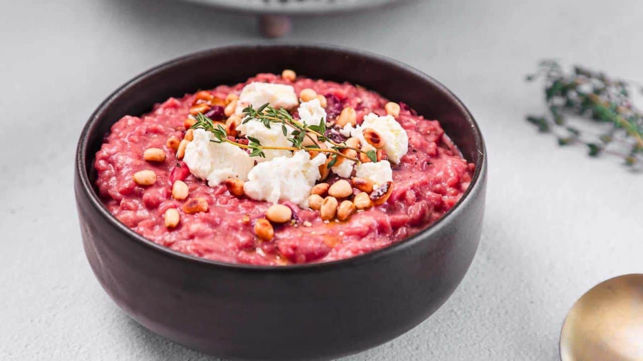 A black bowl filled with beetroot risotto, topped with pine nuts, goat cheese, and a small sprig of thyme, placed on a light-colored surface.