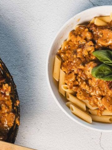 A bowl of penne pasta topped with a creamy, tomato-based meat sauce and basil, beside a skillet with more sauce and a wooden spatula.