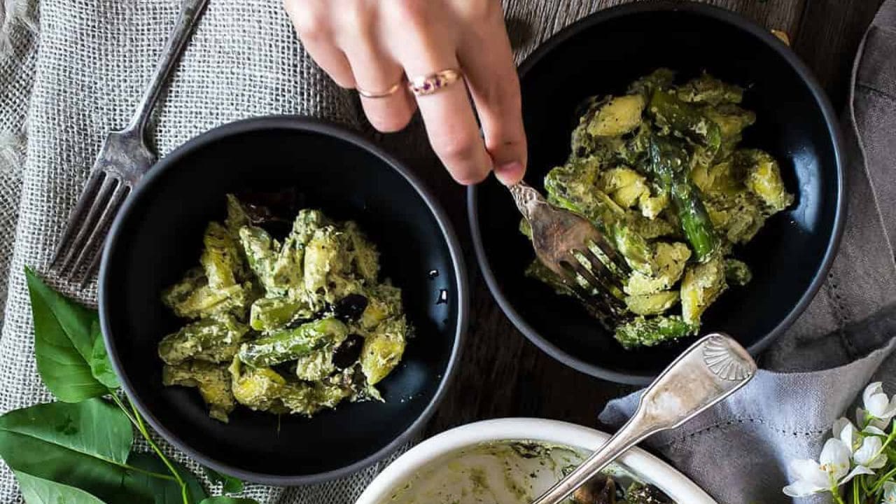A bowl of pasta salad featuring mushrooms, asparagus, and a creamy green sauce. The dish is garnished with fresh herbs. A small bowl of nuts and some greenery are in the blurred background.
