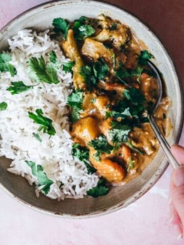A hand holds a spoon over a bowl of white rice and curry garnished with fresh cilantro on a light pink surface.