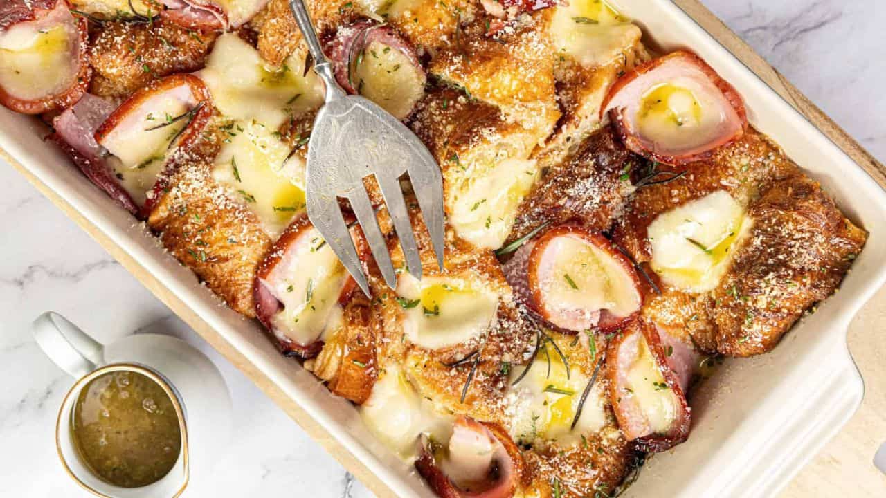A fork lifts a portion of croissant breakfast casserole with cheese and slices of fruit, from a baking dish. A cup sits in the background.