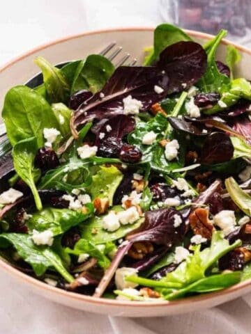 A bowl of mixed green salad with feta cheese, dried cranberries, and pecans, served with two forks on a white surface.