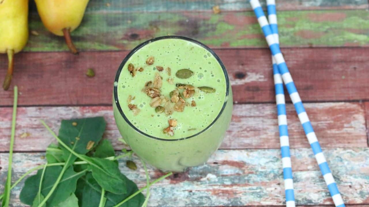 A green smoothie in a glass topped with granola, surrounded by spinach leaves, two pears, and blue-striped straws on a rustic wooden surface.