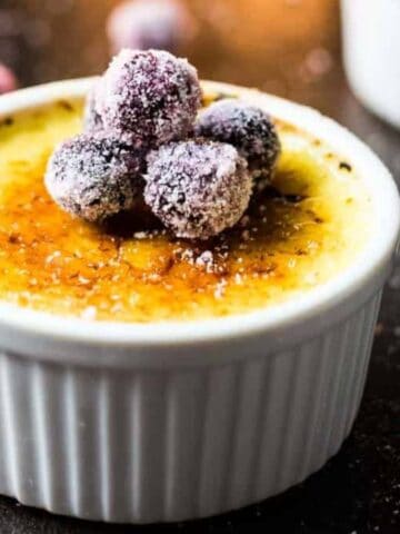 A ramekin of crème brûlée topped with sugared berries sits on a dark surface, with more sugared berries scattered around.