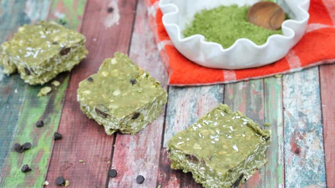 Three matcha oat bars with chocolate chips are placed on a rustic wooden surface, with a bowl of green matcha powder and a hazelnut on an orange cloth in the background.