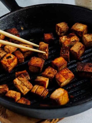 A hand uses chopsticks to pick up a piece of browned tofu from a cast iron skillet with several cooked tofu cubes inside.