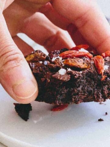 A hand holds a piece of chocolate brownie topped with dried red berries over a white plate.