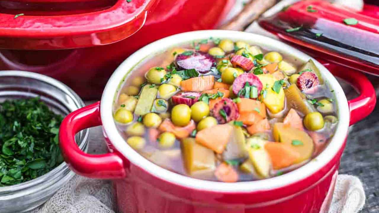 A pot of soup with vegetables and herbs on a wooden table.