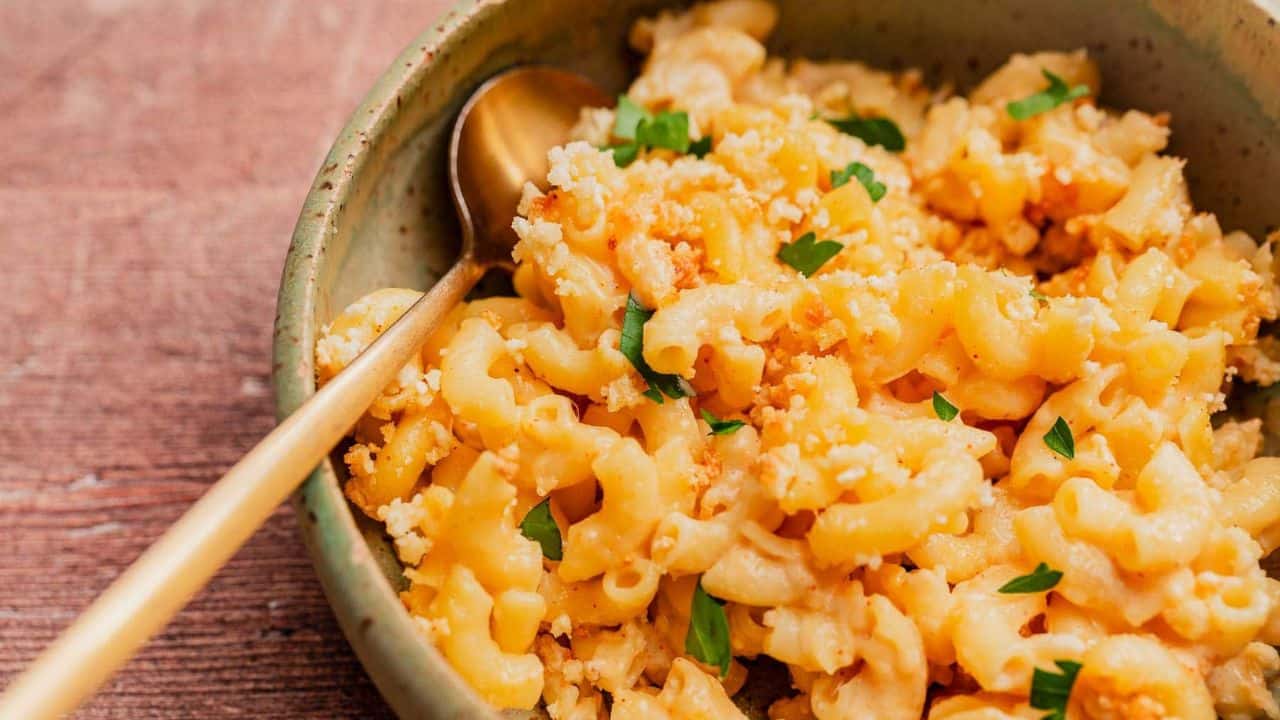 A bowl of macaroni and cheese topped with breadcrumbs and parsley, with a gold spoon resting inside the bowl.