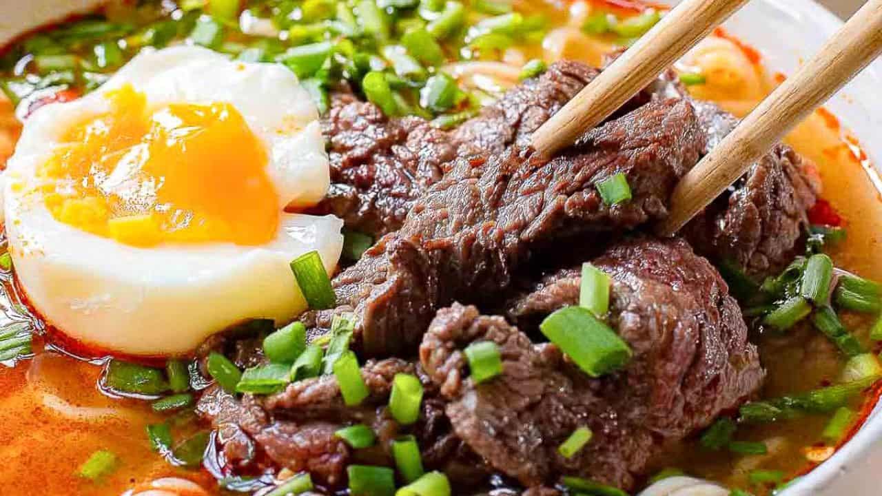 Close-up of a bowl of ramen with rich broth, topped with slices of tender beef, a perfectly poached egg with a runny yolk, and garnished with chopped green onions. Chopsticks are holding up a piece of beef.