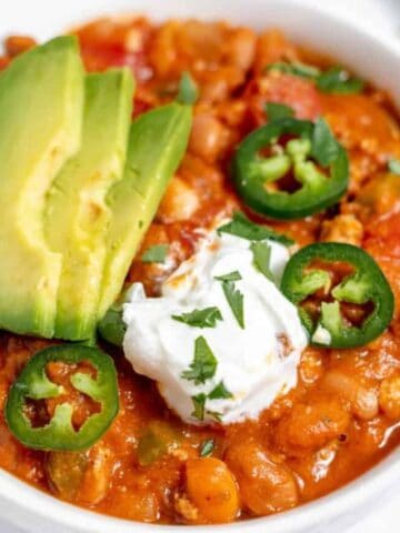 A bowl of chili topped with sliced avocado, jalapeño, a dollop of sour cream, and chopped cilantro, with a spoon on the side.