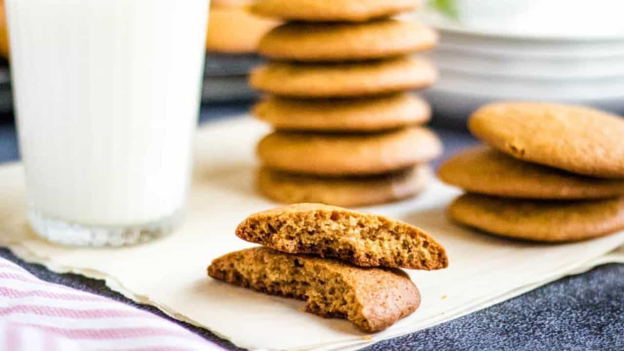 Low-angled shot of a honey cookie broken in half with stacks of honey cookies and a glass of milk in the background.