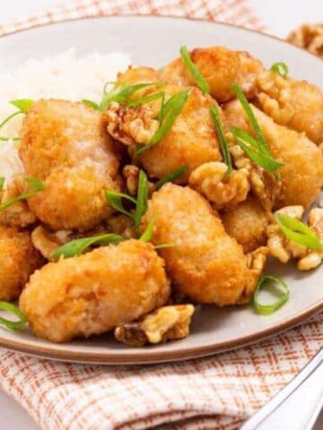 A plate with breaded chicken pieces, white rice, walnut pieces, and sliced green onions, placed on a checkered cloth with a fork beside it.