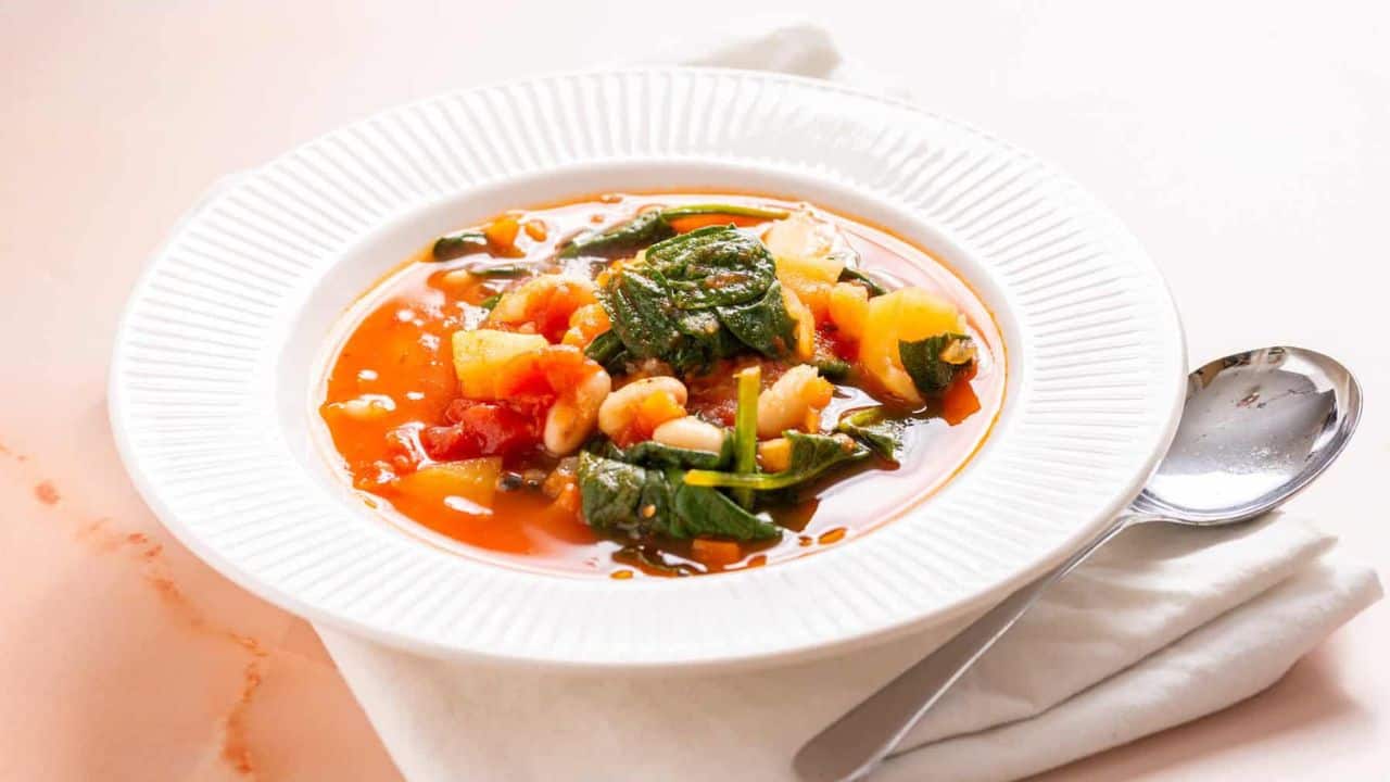 An overhead image of a bowl of minestrone soup served in a white bowl, accompanied by a spoon on the side and a white cloth underneath.