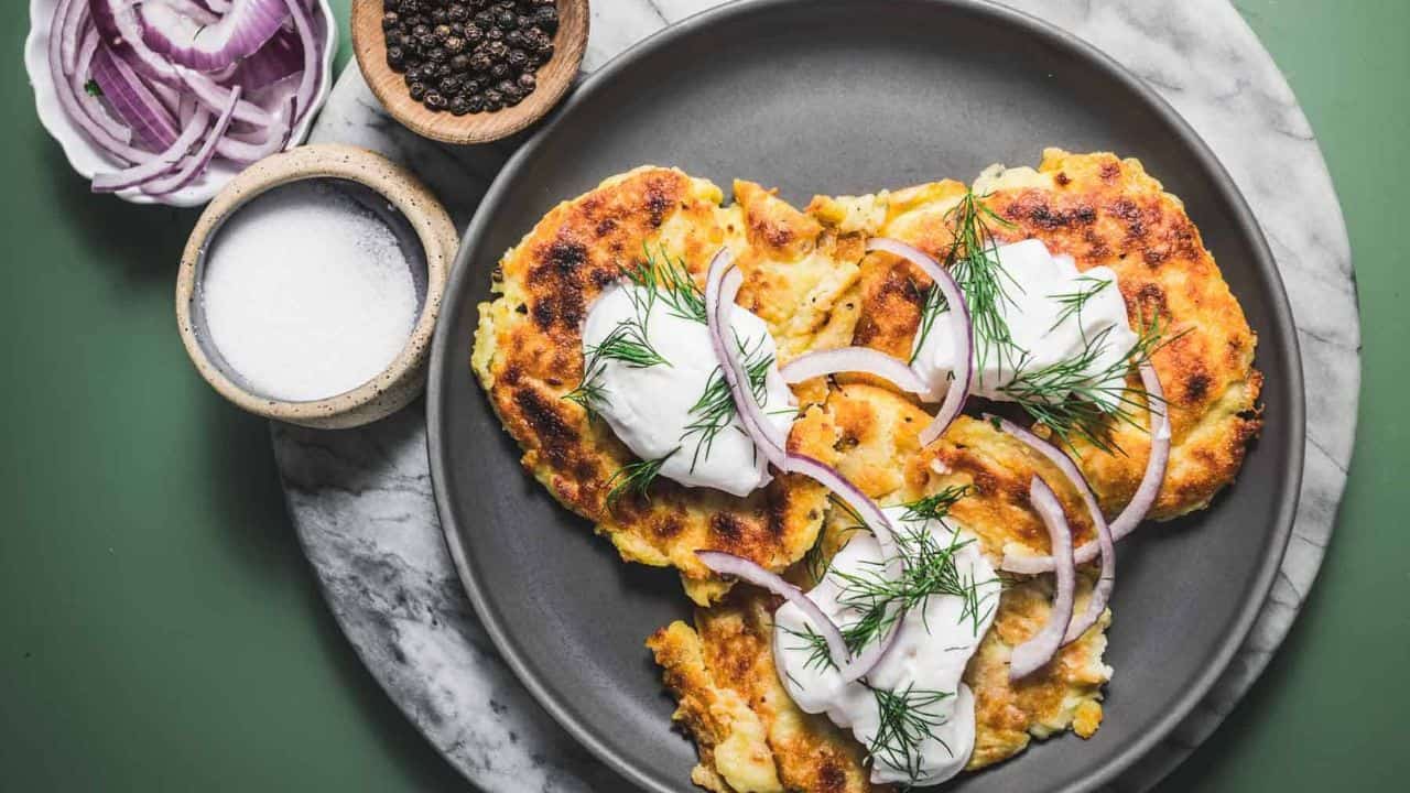 Three potato pancakes topped with sour cream, red onion slices, and dill on a gray plate, with bowls of salt, pepper, and more onions on the side.