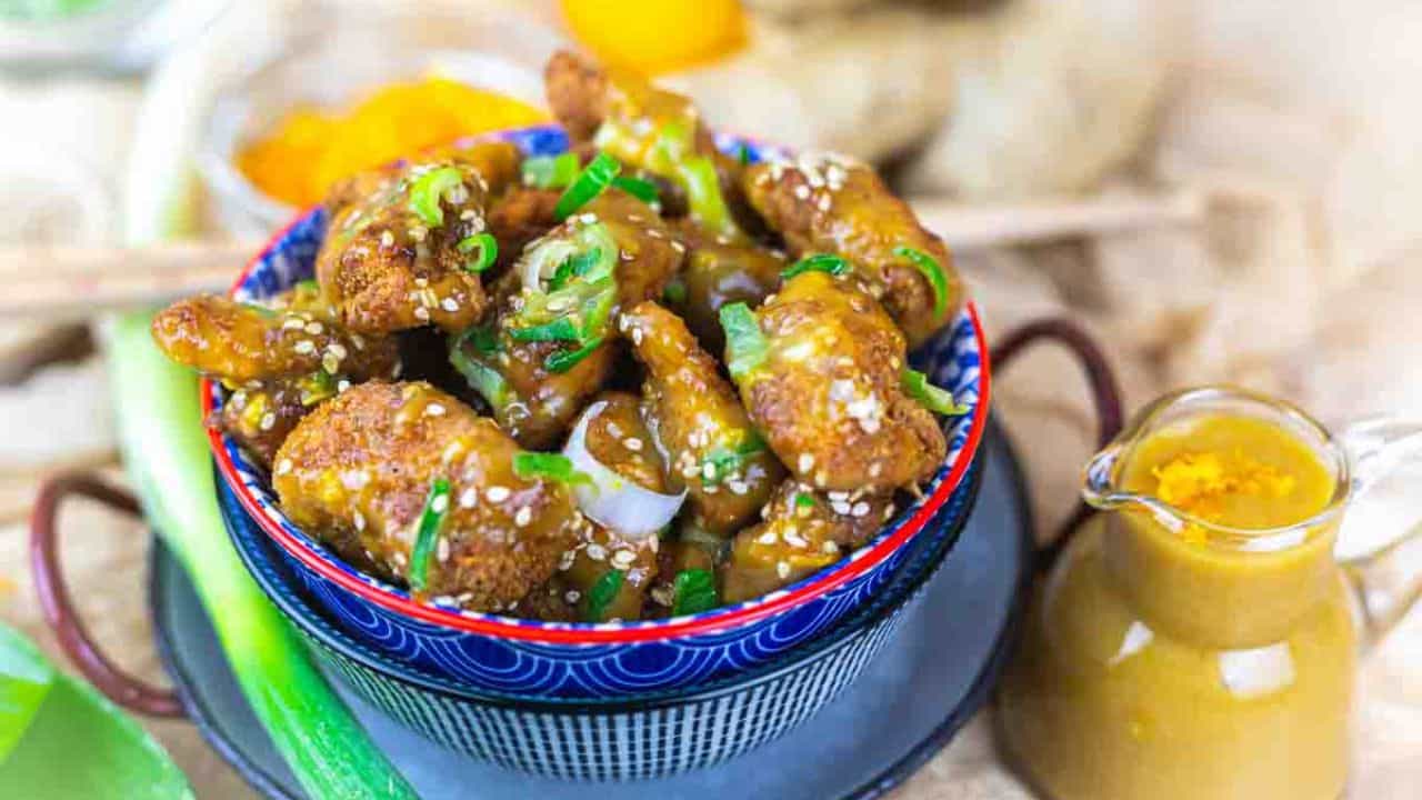 A bowl of fried chicken pieces coated in sauce and garnished with sesame seeds and green onions, accompanied by a small jug of sauce.