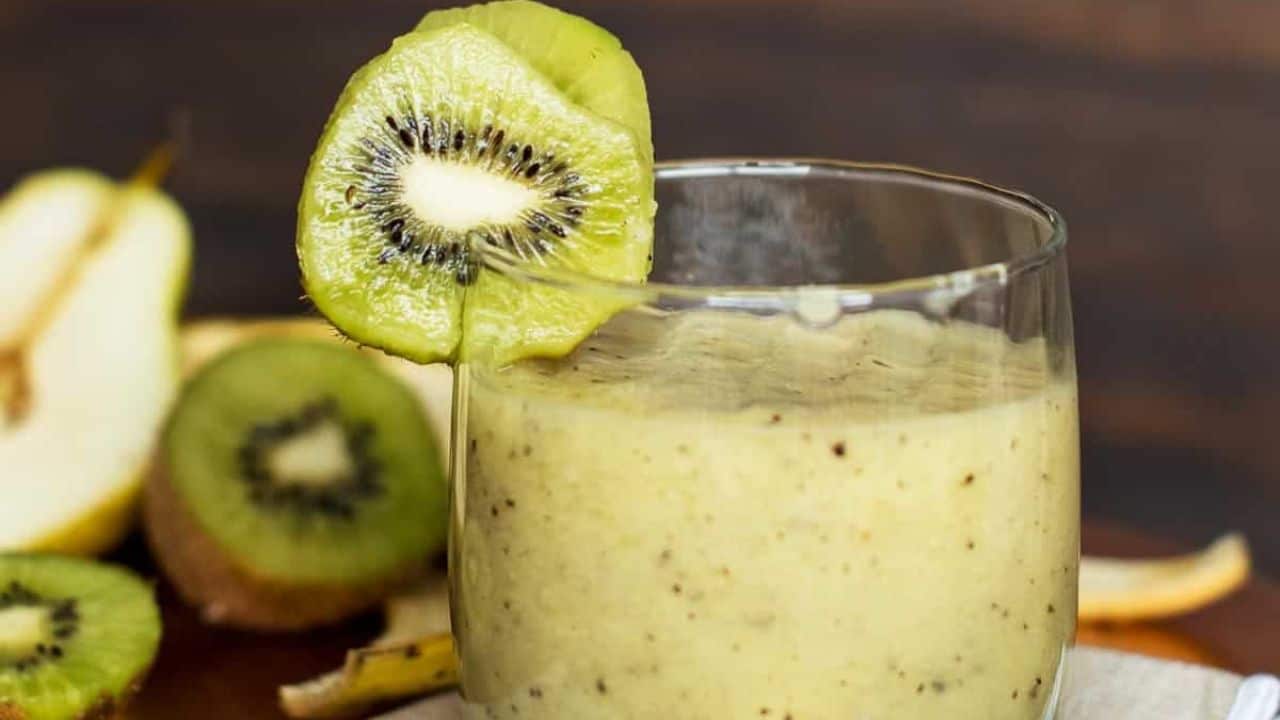 A kiwi pear green smoothie in a glass with a kiwi on the rim and a green straw.