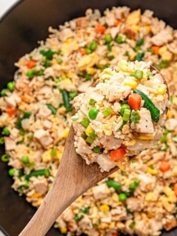 A wooden spoon holds a portion of vegetable and chicken fried rice above a wok filled with the same dish.