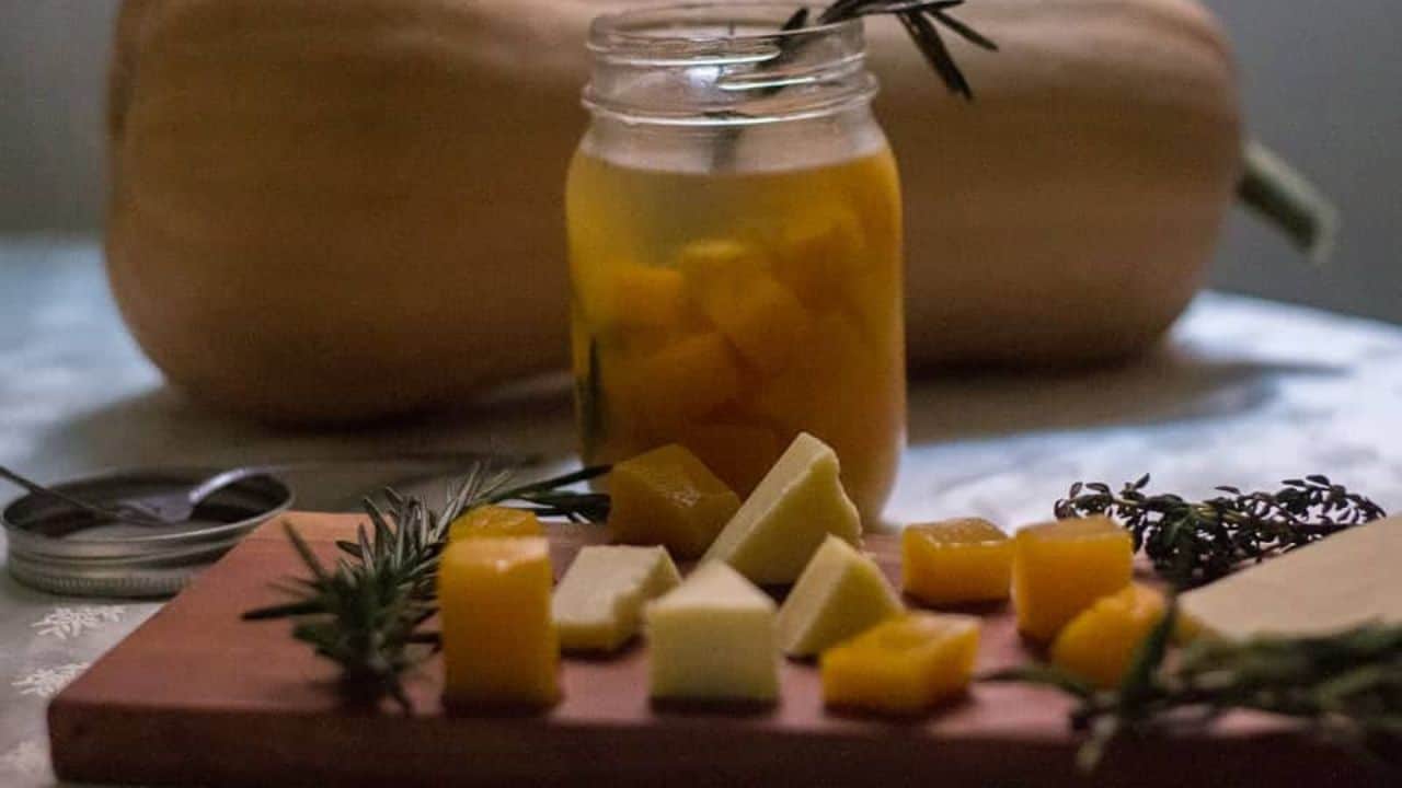 A wooden cutting board with cheese, herbs and a pumpkin.