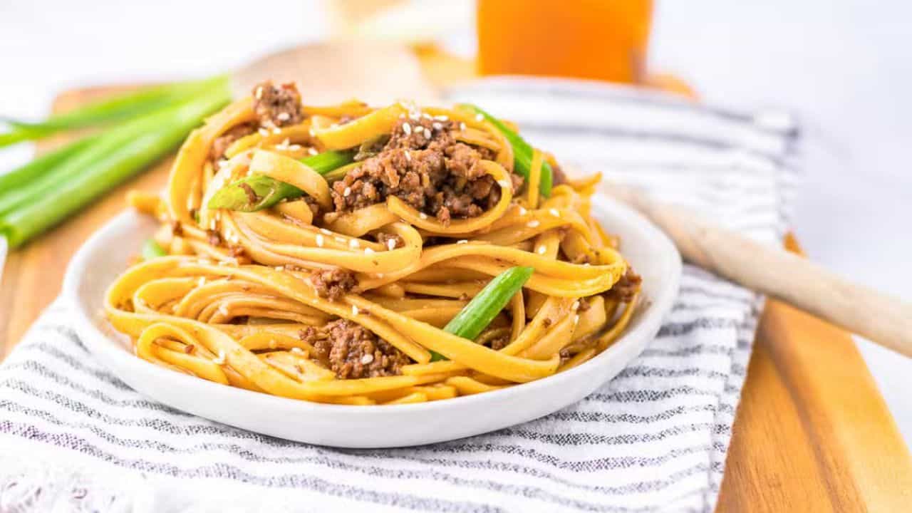 A close-up of a fork holding a bite of pasta with ground meat, green onions, and sauce. The pasta is wrapped around the fork tines, displaying the dish's ingredients and textures in detail, with a blurred background featuring more of the meal.