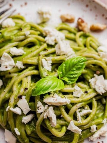 Plate of spaghetti with green pesto sauce, topped with crumbled cheese and walnuts, garnished with a basil leaf, and a fork resting on the side.