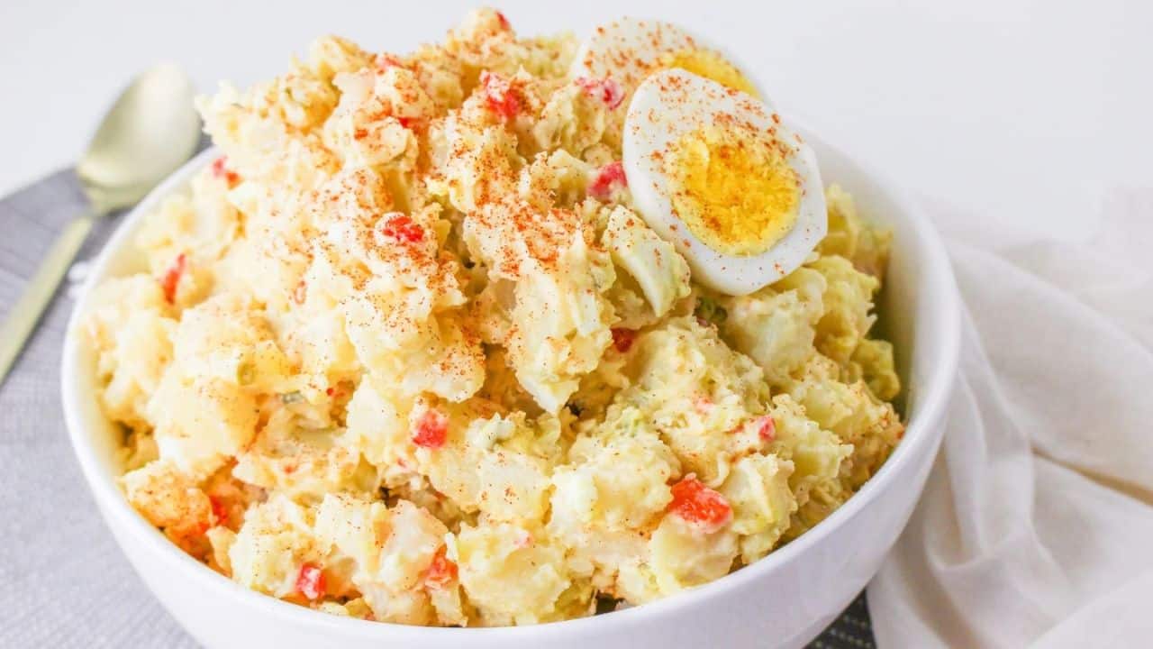 A bowl of potato salad garnished with sliced boiled eggs and a sprinkle of paprika, placed on a table with a white napkin and a gold spoon nearby.