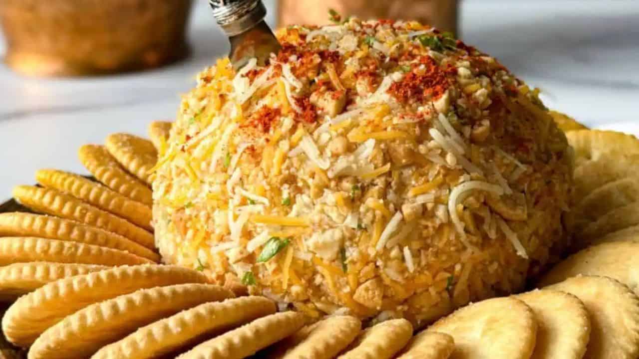The image shows a cheese ball coated with nuts and spices, garnished with parsley. It is surrounded by a ring of round crackers on a platter, with a serving knife inserted into the top.