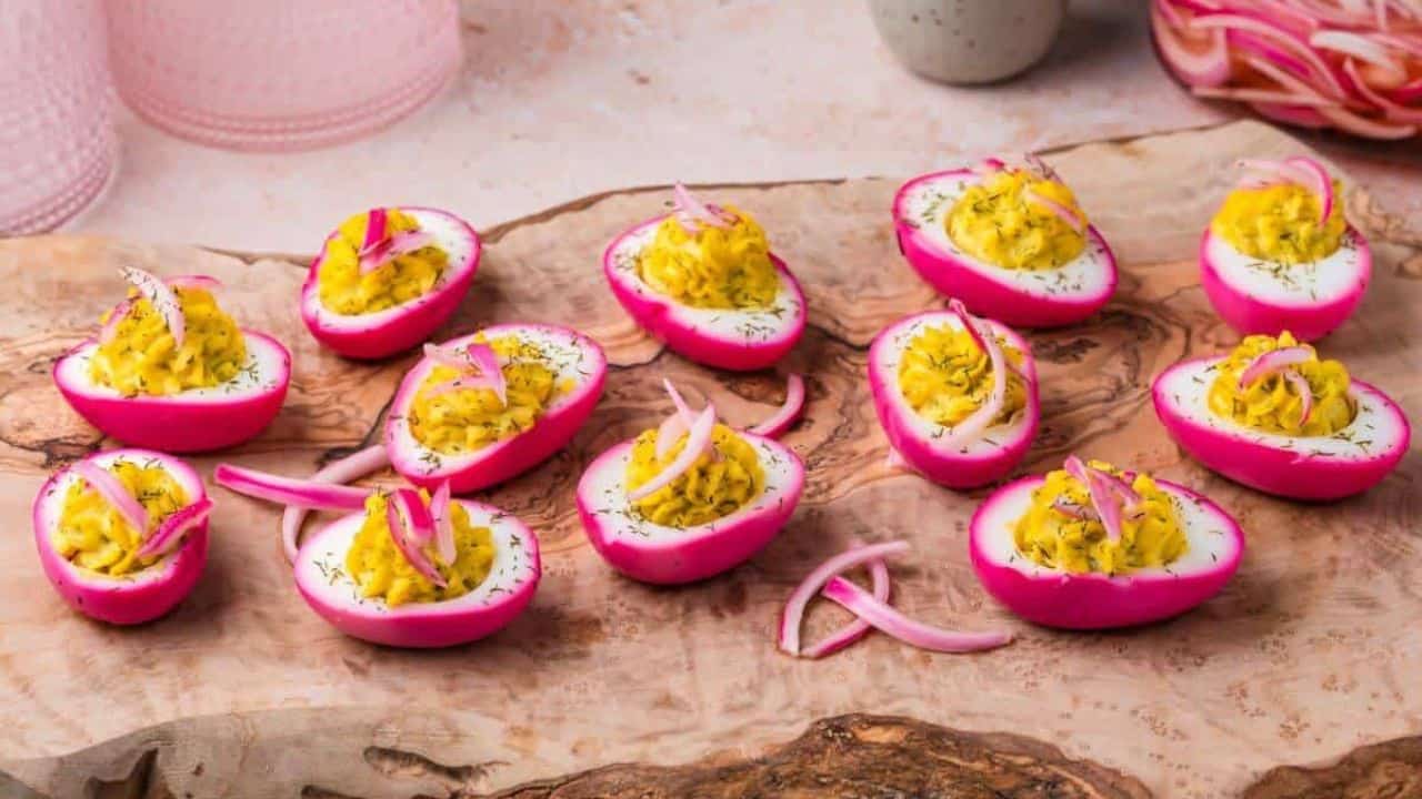 Deviled eggs with bright pink edges and yellow filling topped with sliced onions arranged on a wooden board.