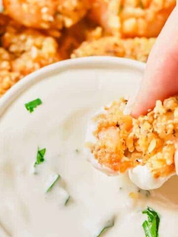 A hand dips a breaded shrimp into a small bowl of white dipping sauce, with more shrimp pieces and chopped herbs visible in the background.