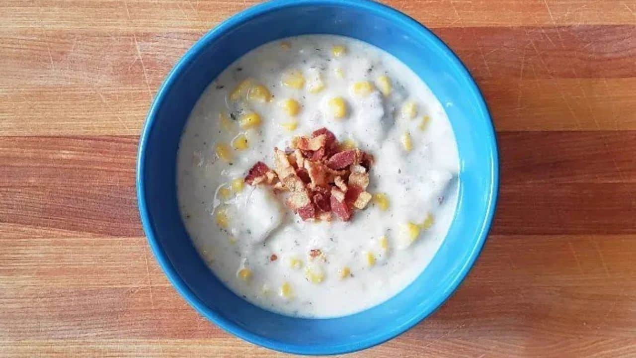 Spoon holding potato and corn chowder with bacon with the full bowl of soup behind it.