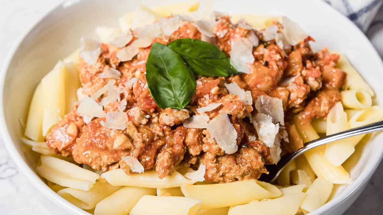 A white bowl filled with penne pasta topped with meat sauce, grated parmesan cheese, and fresh basil leaves, with a metal spoon resting on the side of the bowl.