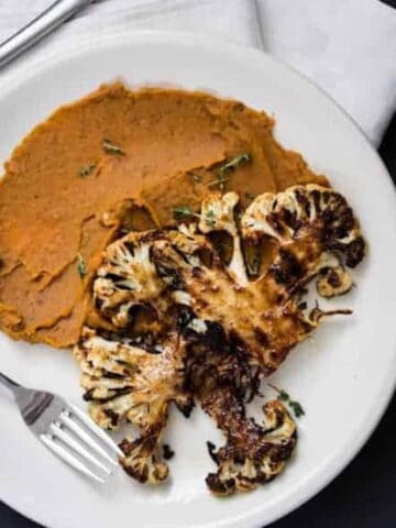 A white plate with two roasted cauliflower steaks and a serving of mashed beans, placed next to a fork and knife, with a glass of light-colored beverage nearby.
