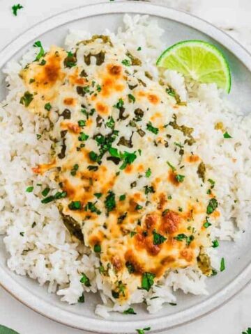 A plate of white rice topped with cheesy green sauce chicken, garnished with chopped herbs, served with a lime wedge. Avocado, lime, and cilantro are nearby.